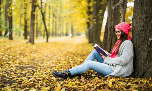 Beautiful young brunette sitting on a fallen autumn leaves in a park, reading a book